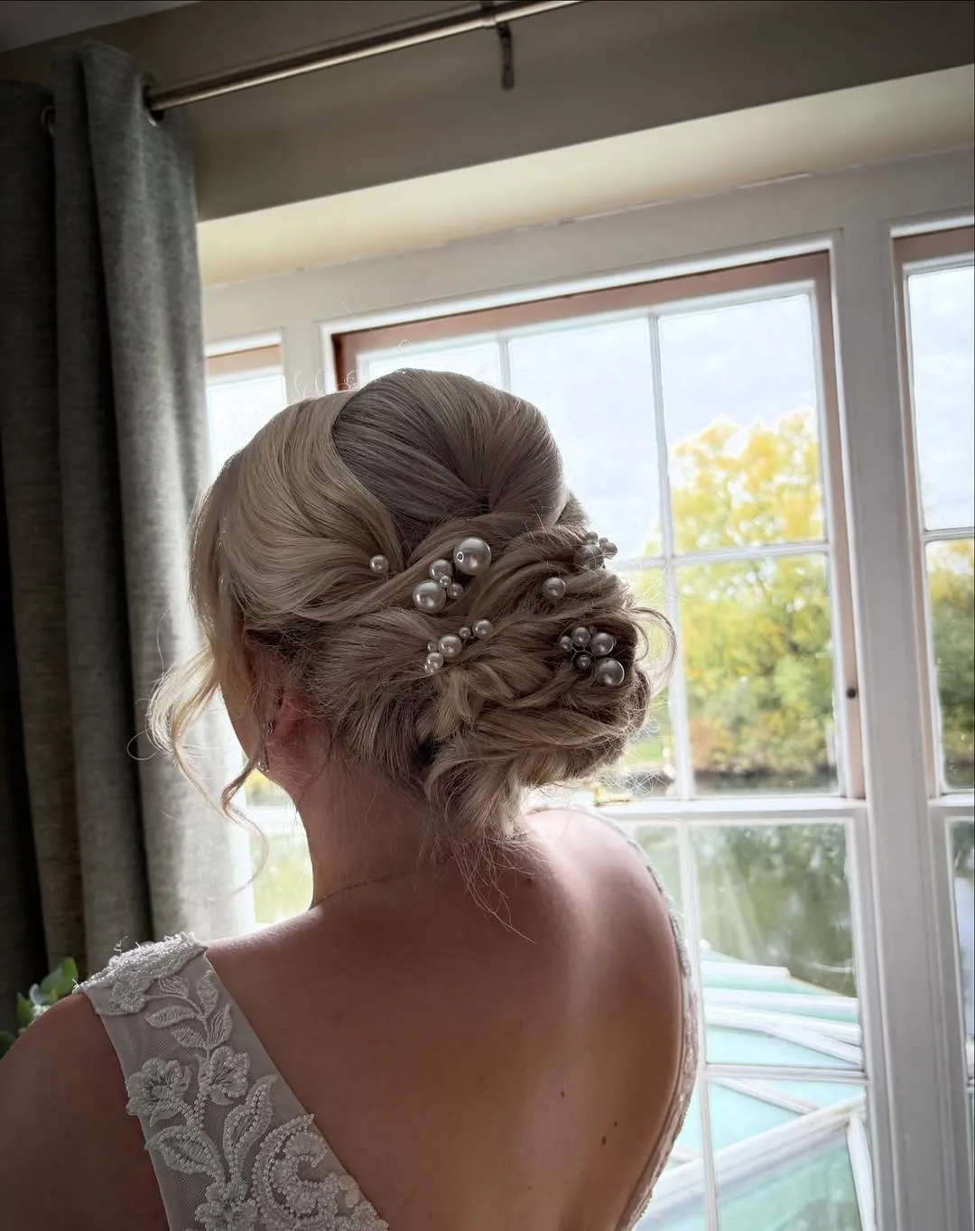 Low, textured blonde chignon studded with white pearl pins, on a bride facing a bright window.