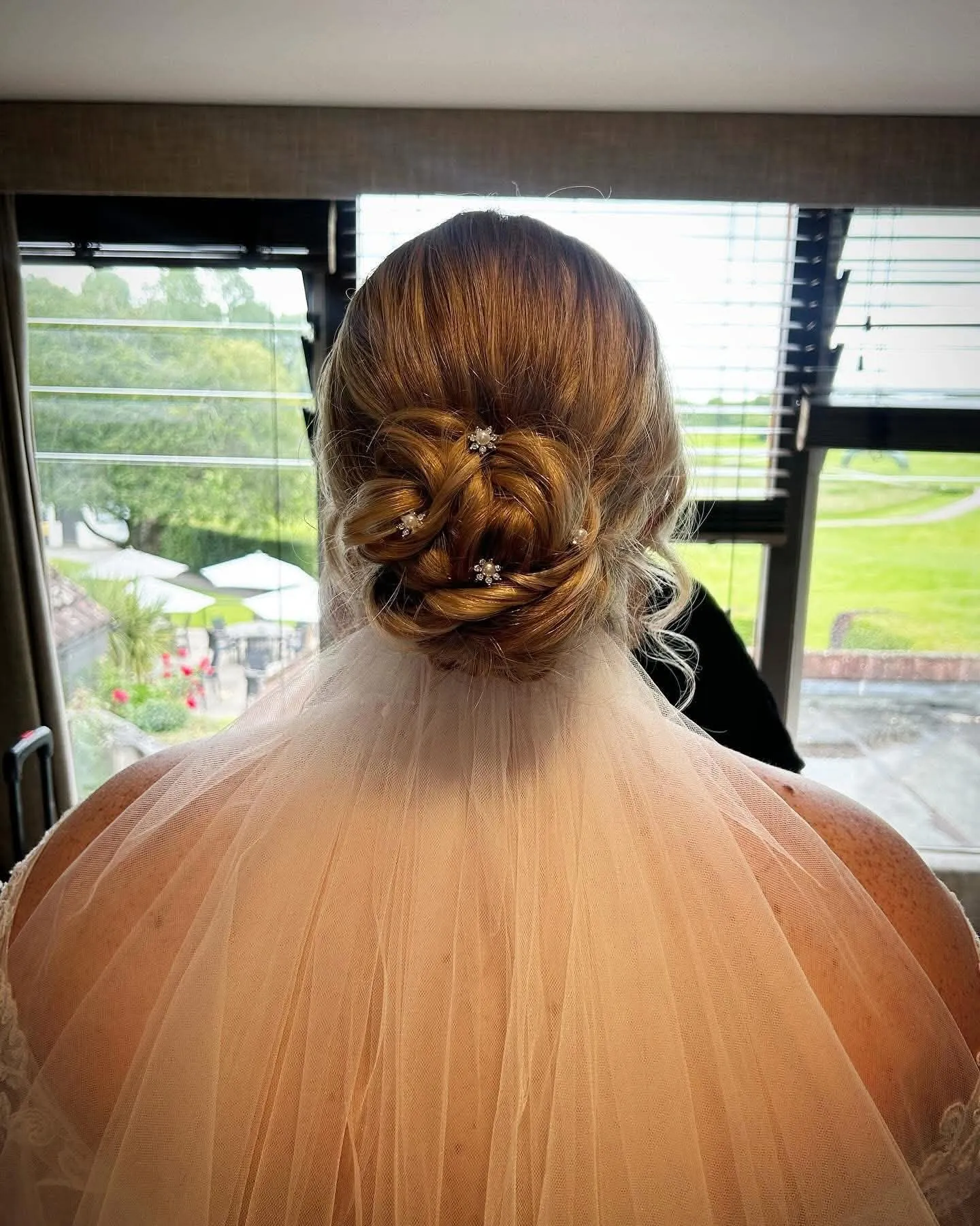 Back view of a bride with a woven low bun and crystal pins, her veil falling from beneath it, by a window